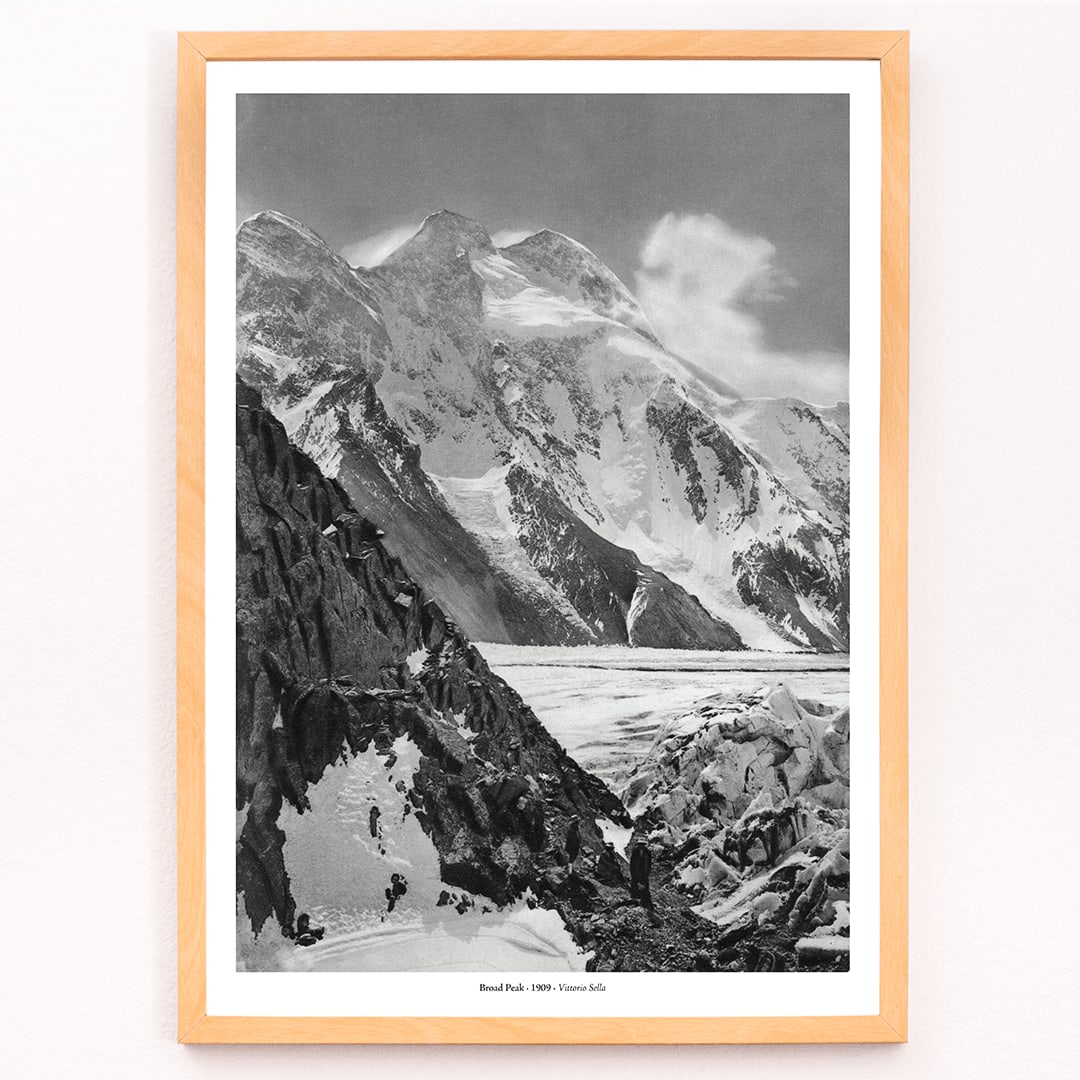 Black and white photograph and poster of Broad Peak by Vittorio Sella showing snow covered mountains and mountaineers climbing amidst rocky terrain and glacier, framed and displayed in modern living room decor