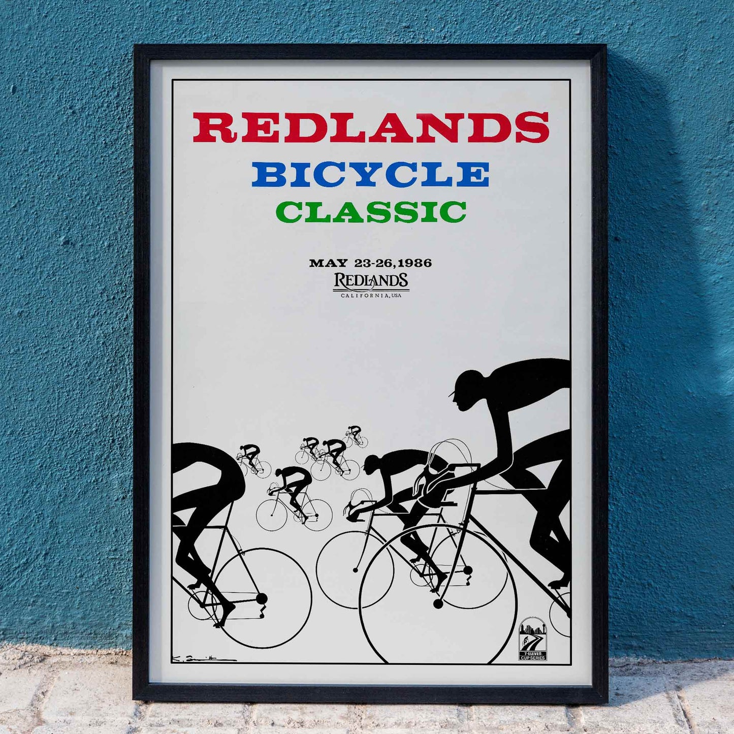 Cyclists racing illustration and poster by Karlis Smiltens titled Redlands Bicycle Classic featuring red blue and green text on white background, framed and displayed in home decor