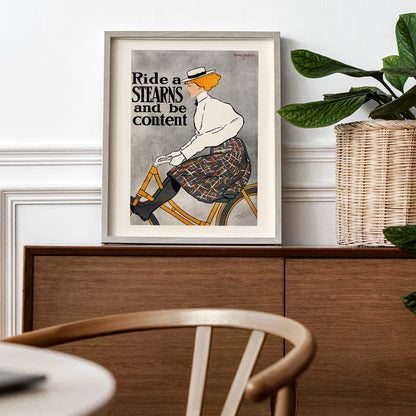 Vintage poster of a woman riding a bicycle with the text Ride a Stearns and be content by Edward Penfield artwork, framed and displayed in home decor